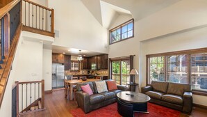 A spacious living area with a high ceiling, a staircase, a kitchen, and large windows.