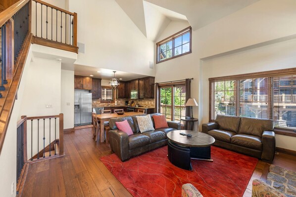 A spacious living area with a high ceiling, a staircase, a kitchen, and large windows.