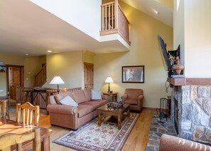 A spacious living room with a high ceiling, a fireplace, a staircase, and a dining area.