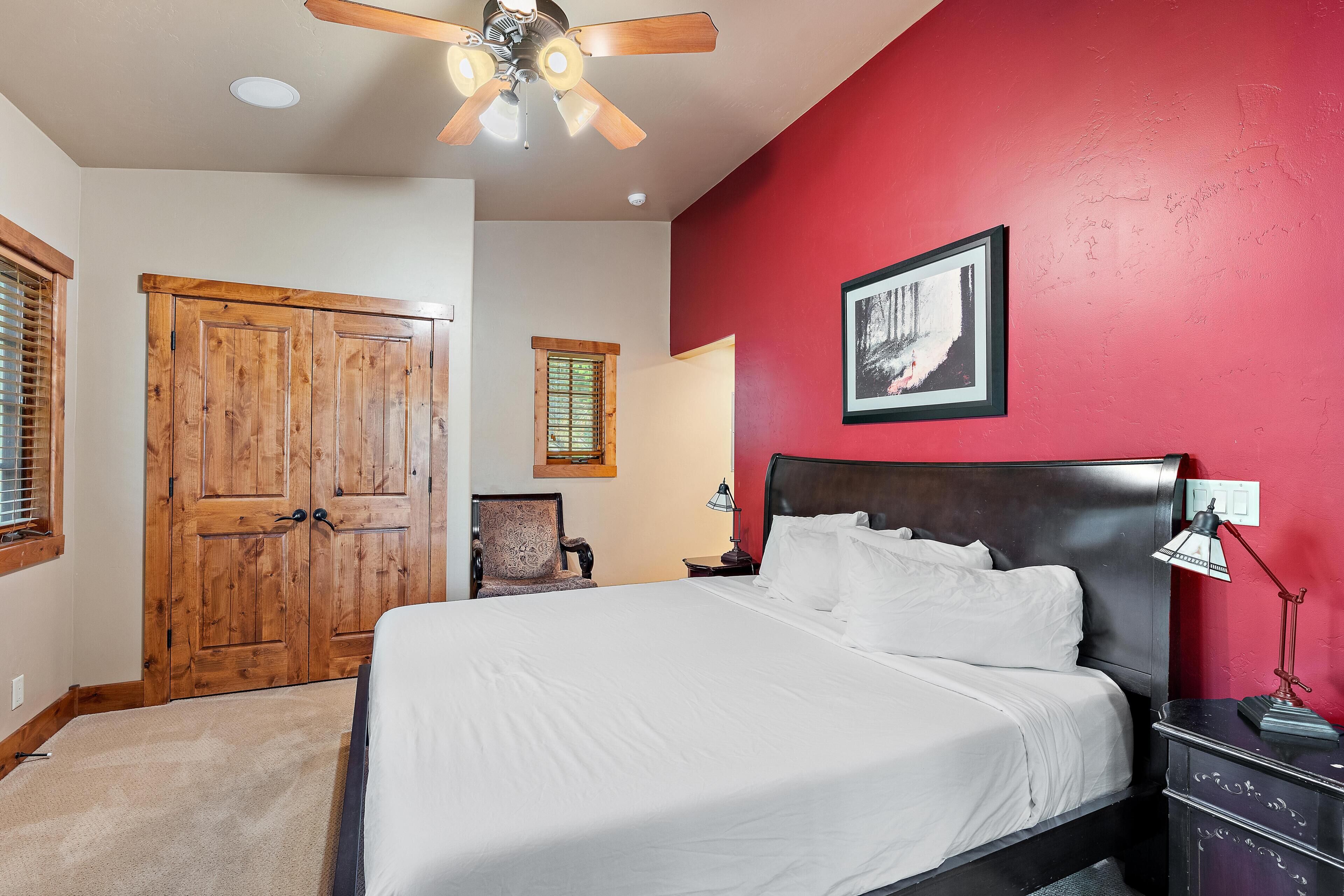 A bedroom with a bed, wooden wardrobe, a ceiling fan, a framed picture on the wall, and a nightstand with a lamp.