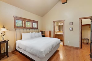 A bedroom with a large bed, wooden headboard, a nightstand, a lamp, a dresser, and a window with a view of trees. - Wonderful Cottage |Ski In/Out | Private Hot Tub (Tamarack)