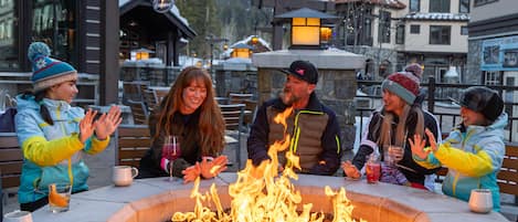 A group of people seated around a fire pit in an outdoor setting with buildings in the background.