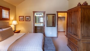Un dormitorio con una cama grande, un armario de madera, un banco, una mesita de noche, una lámpara y un espejo.