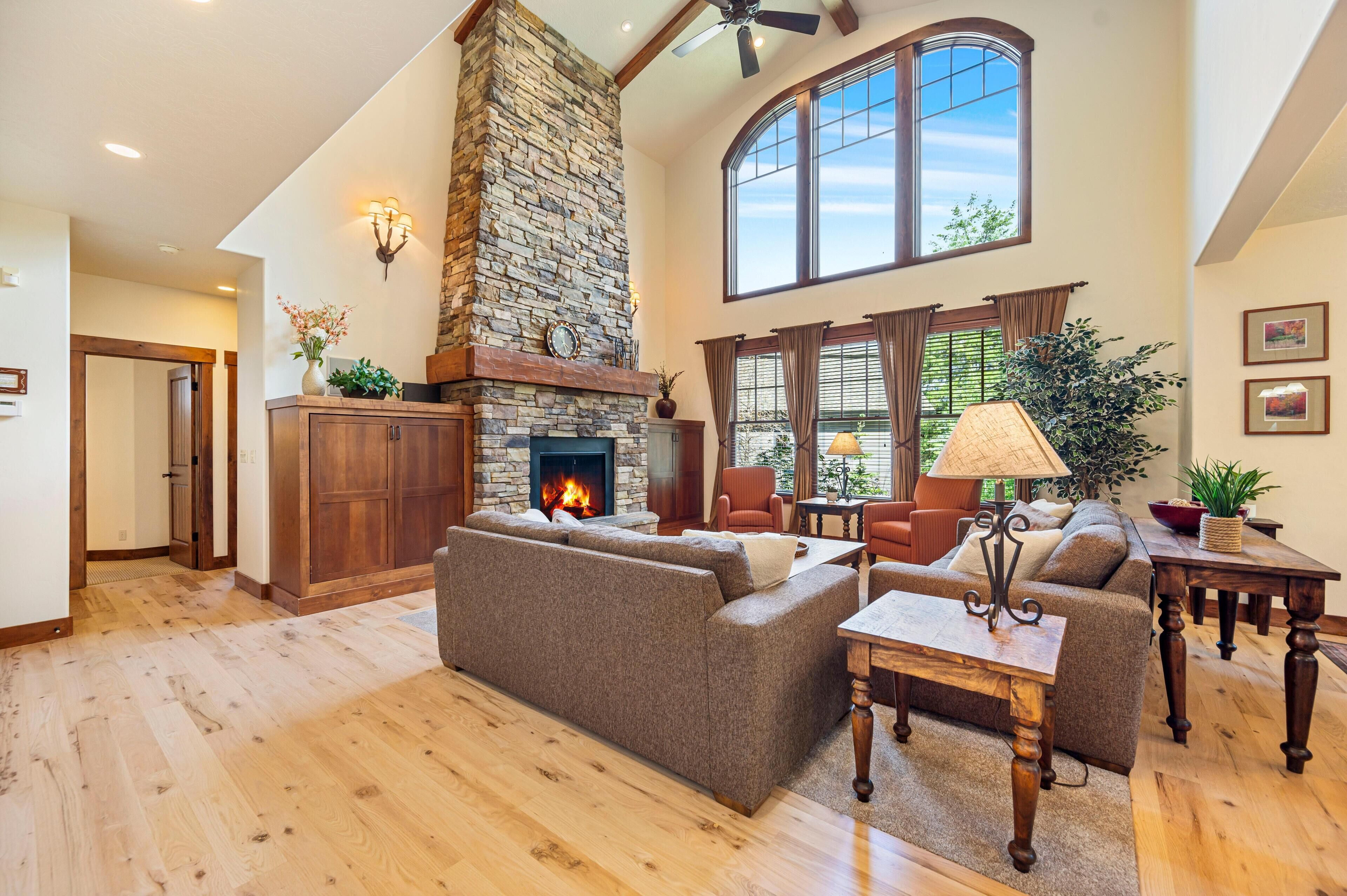 A spacious living room with a stone fireplace, wooden flooring, and large windows.