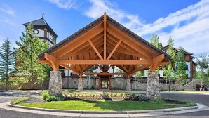 A wooden pavilion with a stone base, surrounded by a landscaped area with trees and a building with a clock tower in the background. - Unique King Studio  with Jet Tub (Tamarack)