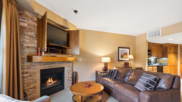 A living room with a fireplace, a sofa, a coffee table, and a TV mounted above the fireplace. - Unique King Studio  with Jet Tub (Tamarack)