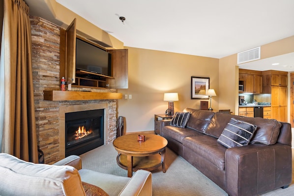 A living room with a fireplace, a sofa, a coffee table, and a TV mounted above the fireplace. - Unique King Studio with Jet Tub (Tamarack)