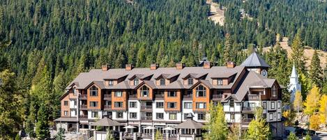 Un hotel de varios pisos con fachada de piedra, varios balcones y un complejo de esquí al fondo.