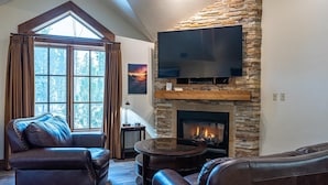 A cozy living room with a stone fireplace, leather furniture, a flat-screen TV, and a large window with curtains. - No Regrets | Exclusive Tamarack Perks (Tamarack)