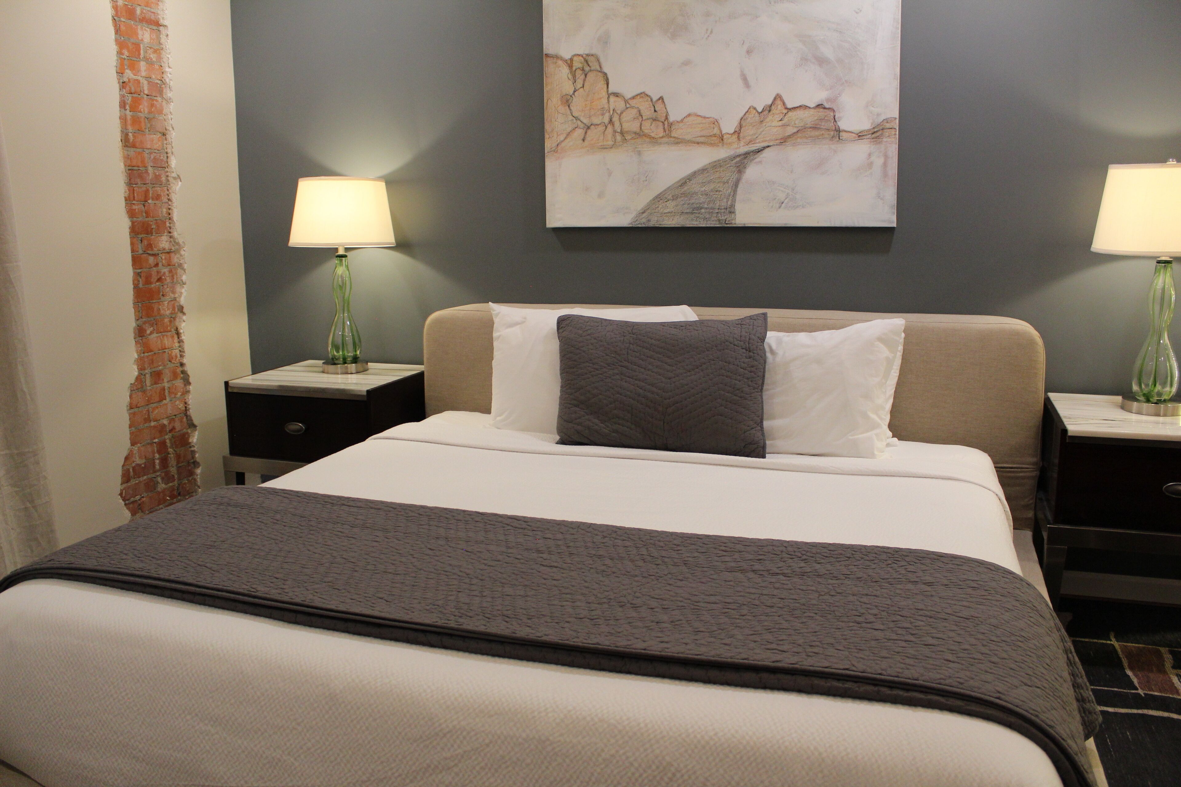 A hotel room with a bed, two bedside tables, lamps, a painting on the wall, and a brick wall feature.
