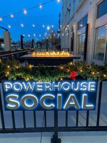 Letrero de 'POWERHOUSE SOCIAL' con contorno de neón, rodeado de luces festivas y follaje.