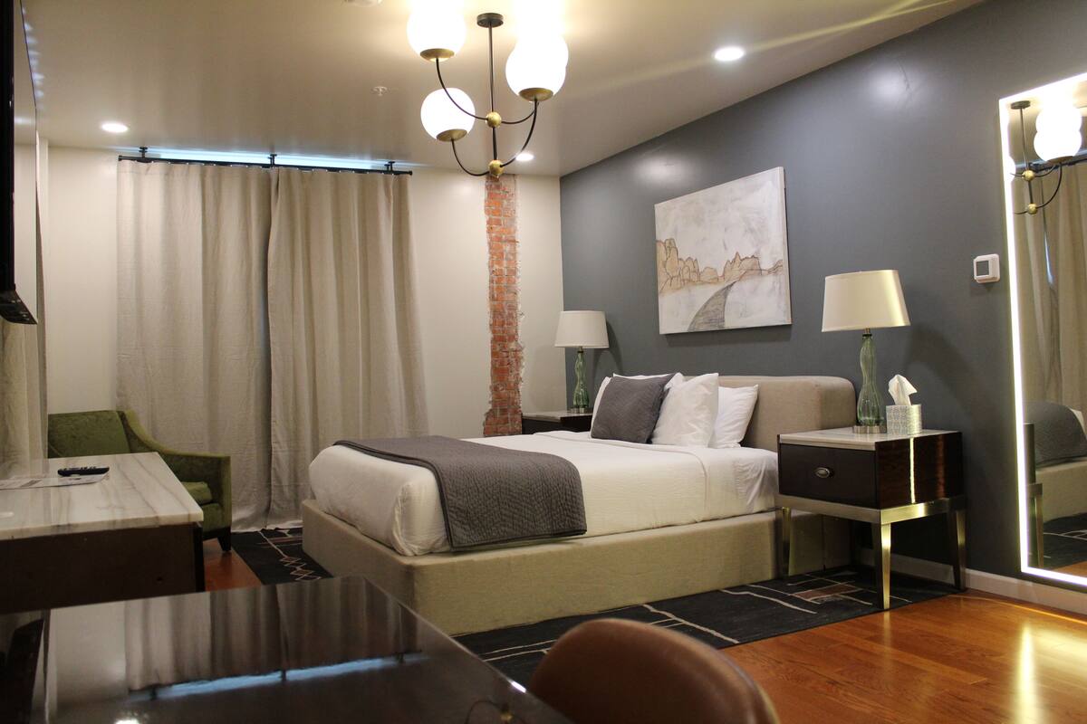 A hotel room with a bed, bedside tables, a desk, a chair, a mirror, and a chandelier.
