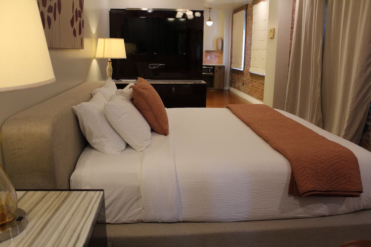 A hotel room with a large bed, a flat-screen TV, a bedside table with a lamp, and a view of the city through the window.