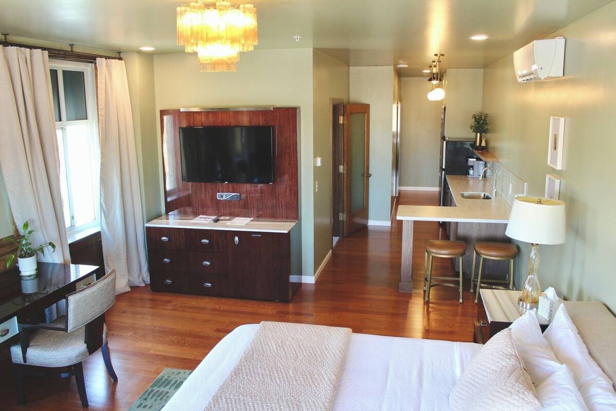 A hotel room with a bed, a desk with a chair, a TV mounted on the wall, a kitchenette, and a bathroom visible through an open door.