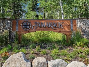 Ein Holzschild mit dem Schriftzug 'Tamarack Resort' und einem Logo, vor einer Steinmauer mit Grünpflanzen im Hintergrund.