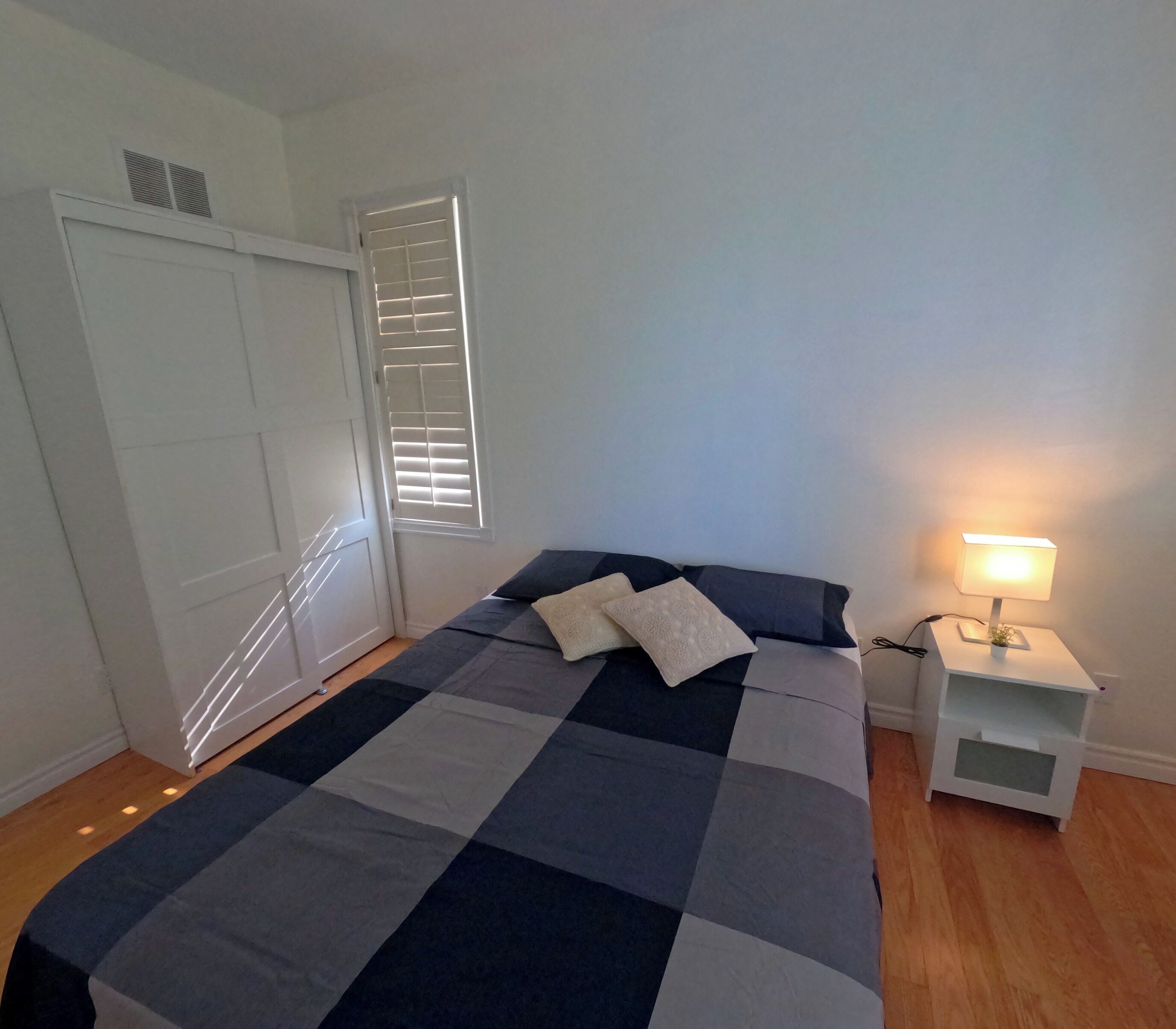 Una camera da letto con un letto, cuscini, un comodino con una lampada, un armadio e una finestra con persiane.