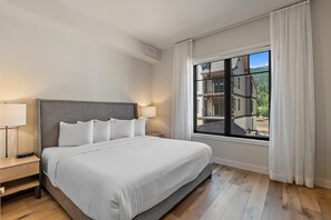 Una cama bien hecha con sábanas blancas, una mesita de noche de madera con una lámpara, y un ventanal con cortinas.