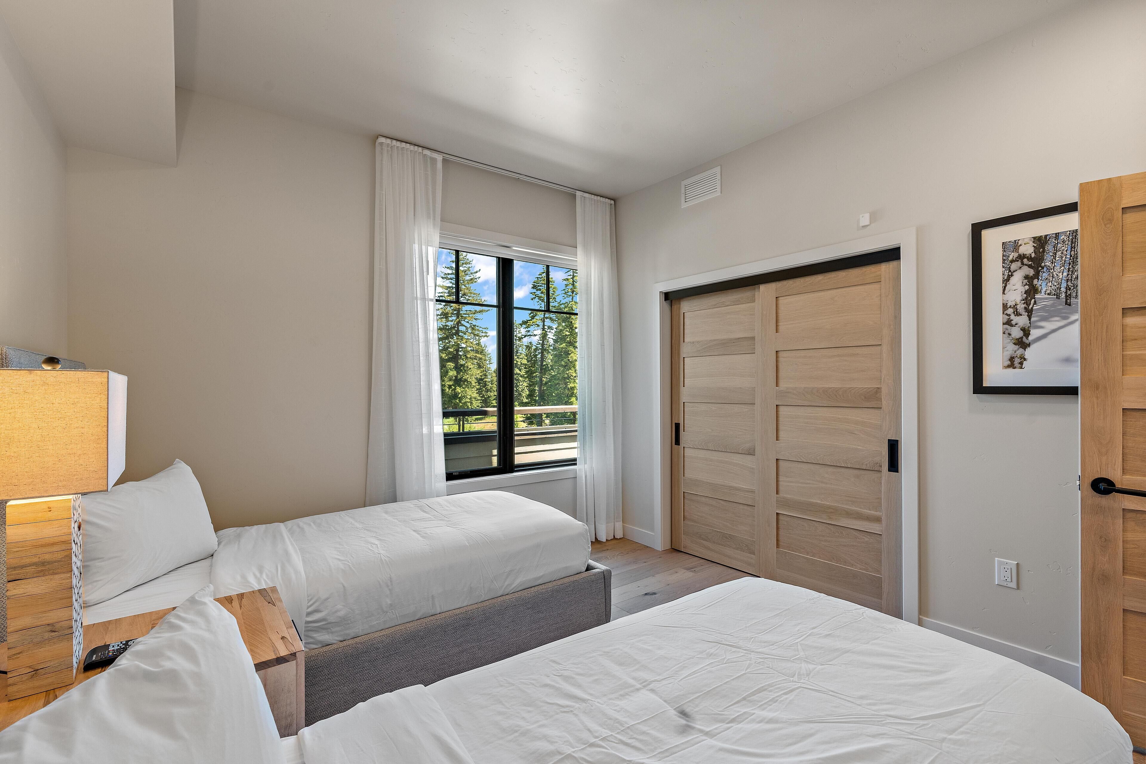 A bedroom with two beds, a window with curtains, a wooden door, and a framed picture on the wall.