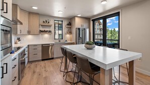 Una cucina moderna con isola centrale, elettrodomestici in acciaio inossidabile, mobili in legno e un'ampia finestra che offre vista su un edificio e alberi.