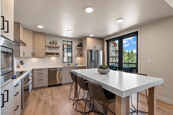 Une cuisine moderne dotée d’un îlot central, d  ’appareils électroménagers en acier inoxydable, de meubles en bois et d’une grande fenêtre offrant une vue sur un immeuble et des arbres.