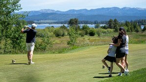 A golf course with a player preparing to hit a ball, a caddie, and a woman walking beside them.