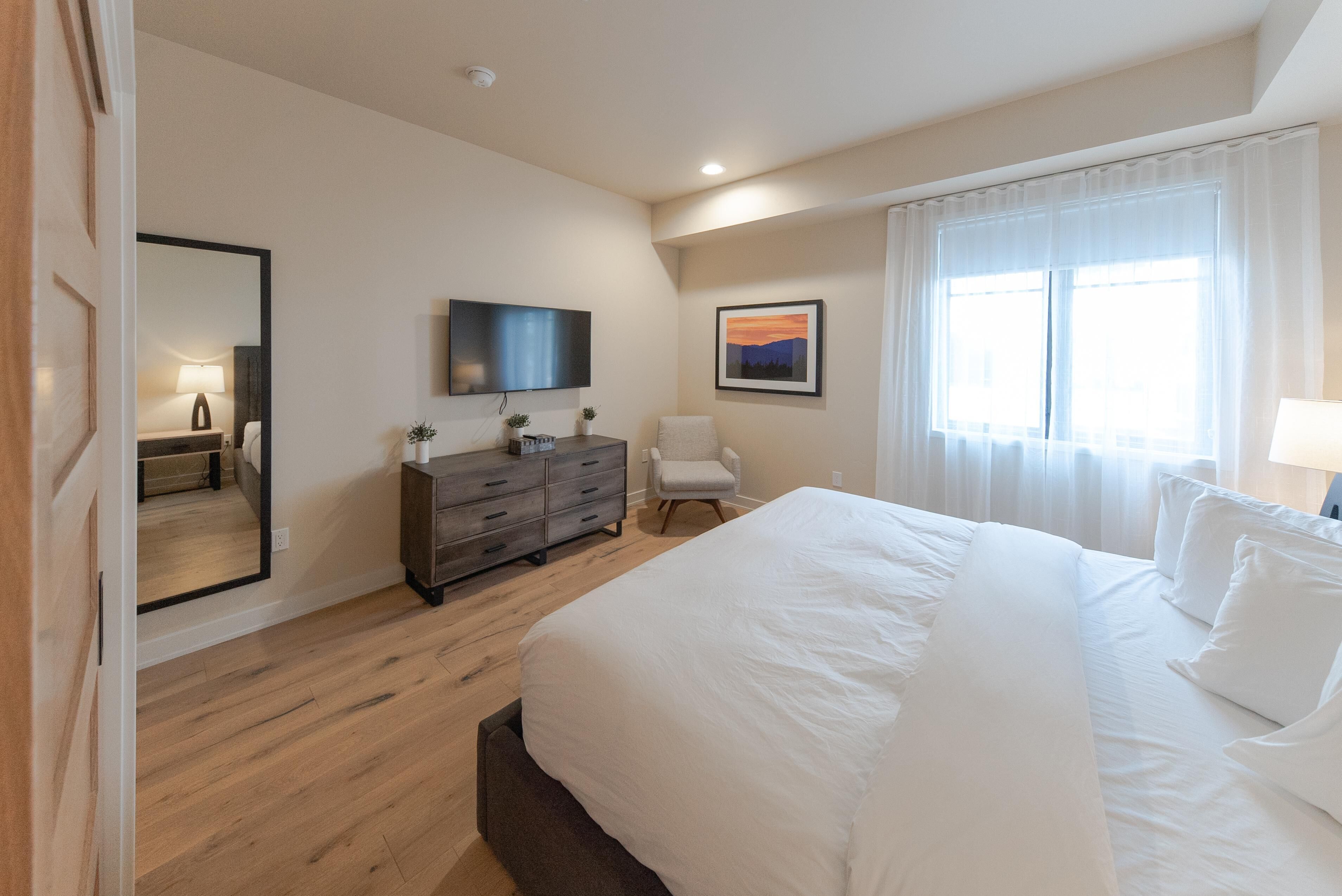 Une chambre à coucher moderne avec un grand lit, une commode, une chaise, un téléviseur et un tableau accroché au mur.