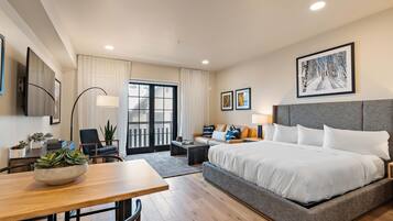 A hotel room with a large bed, a desk, a chair, a sofa, a dining table, and a view of the outside.