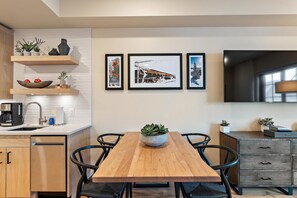 A modern kitchen with wooden cabinets, a dining table, and black chairs. There are three framed pictures on the wall, a coffee machine, and a potted plant on the counter.
