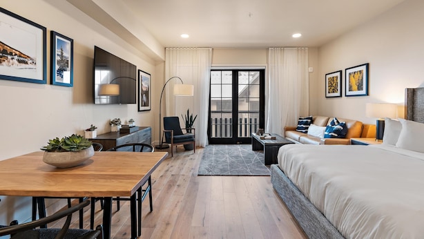 A modern hotel room with a wooden dining table, a sofa, a bed, and a balcony with curtains. - Gorgeous Condo|Juliette Balcony|Mountain & Village Views (Tamarack)
