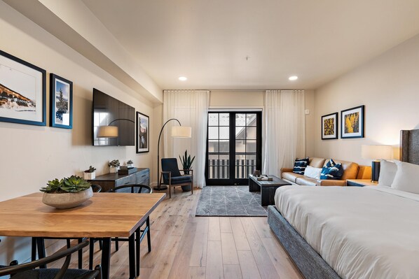Une chambre d’hôtel moderne avec une table à manger en bois, un canapé, un lit et un balcon agrémenté de rideaux.