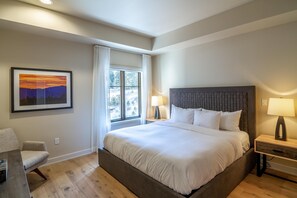 Une chambre avec un grand lit, une fenêtre avec des rideaux, une table de chevet avec une lampe et un tableau encadré au mur.