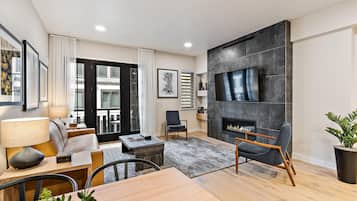 Een moderne woonkamer met een open haard, een flatscreen televisie aan de muur, een eethoek met een tafel en stoelen, en een groot raam met gordijnen.