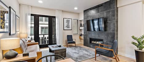 A modern living room with a fireplace, a flat-screen TV mounted on the wall, a dining area with a table and chairs, and a large window with curtains.