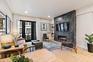 Un salon moderne doté d’une cheminée, d’un téléviseur à écran plat fixé au mur, d’un coin repas avec une table et des chaises, et d’une grande fenêtre agrémentée de rideaux.