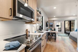 A modern kitchen with stainless steel appliances, a dining area with a wooden table, and a view of the living room through an open door.