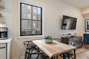 A modern kitchen with a large window, a coffee machine, a dining table, chairs, a TV mounted on the wall, and a small potted plant.