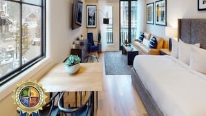 Ein kleines Hotelzimmer mit einem Bett, einem Schreibtisch mit Stuhl, einer Couch und einem großen Fenster mit Blick auf umliegende Gebäude.