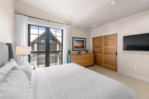 Un dormitorio con una cama grande, un aparador, un televisor montado en la pared y un balcón con vista a una casa.