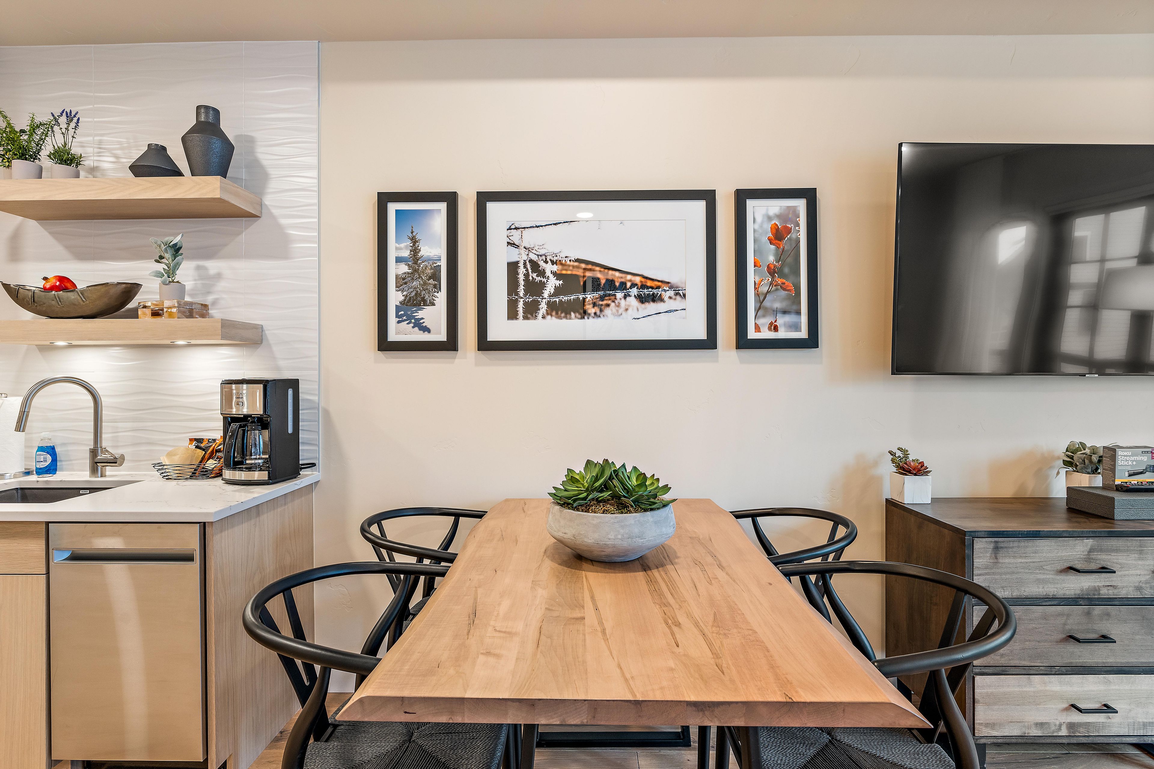 A modern kitchen with a wooden dining table, black chairs, a coffee machine, and a TV mounted on the wall.