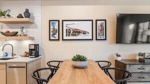Une cuisine moderne avec une table à manger en bois, des chaises noires, une machine à café et un téléviseur fixé au mur.