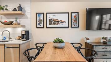 A modern kitchen with a wooden dining table, black chairs, a coffee machine, and a TV mounted on the wall.