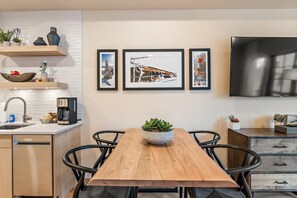 A modern kitchen with a wooden dining table, black chairs, a coffee machine, and a TV mounted on the wall. - Impeccable Condo|Pet Friendly|Juliette Balcony (Tamarack)