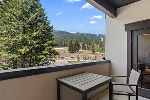 A balcony with a table and chair, overlooking a road with vehicles and a forested hillside.