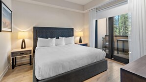 A modern hotel room with a large bed, two bedside tables, a desk, and a balcony with a view of trees.