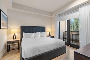 Habitación de hotel moderna con una cama grande, dos mesitas de noche, un escritorio y un balcón con vistas a árboles.