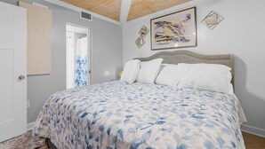 Une chambre à coucher comprenant un lit, un ventilateur de plafond, une porte et un mur orné d’un tableau abstrait encadré.