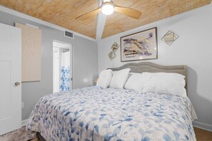 Une chambre à coucher comprenant un lit, un ventilateur de plafond, une porte et un mur orné d’un tableau abstrait encadré.
