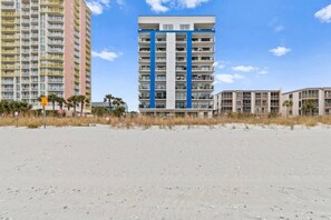 A beachfront hotel with multiple-story buildings, a sandy beach in the foreground, and a clear sky.