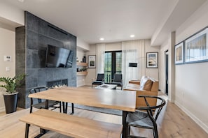 Een moderne woonkamer met een houten eettafel, stoelen, een bank en een televisie die aan een gestructureerde muur is bevestigd.