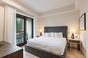 A hotel room with a large bed, two bedside tables with lamps, a view through a sliding glass door, and a small desk with a plant.
